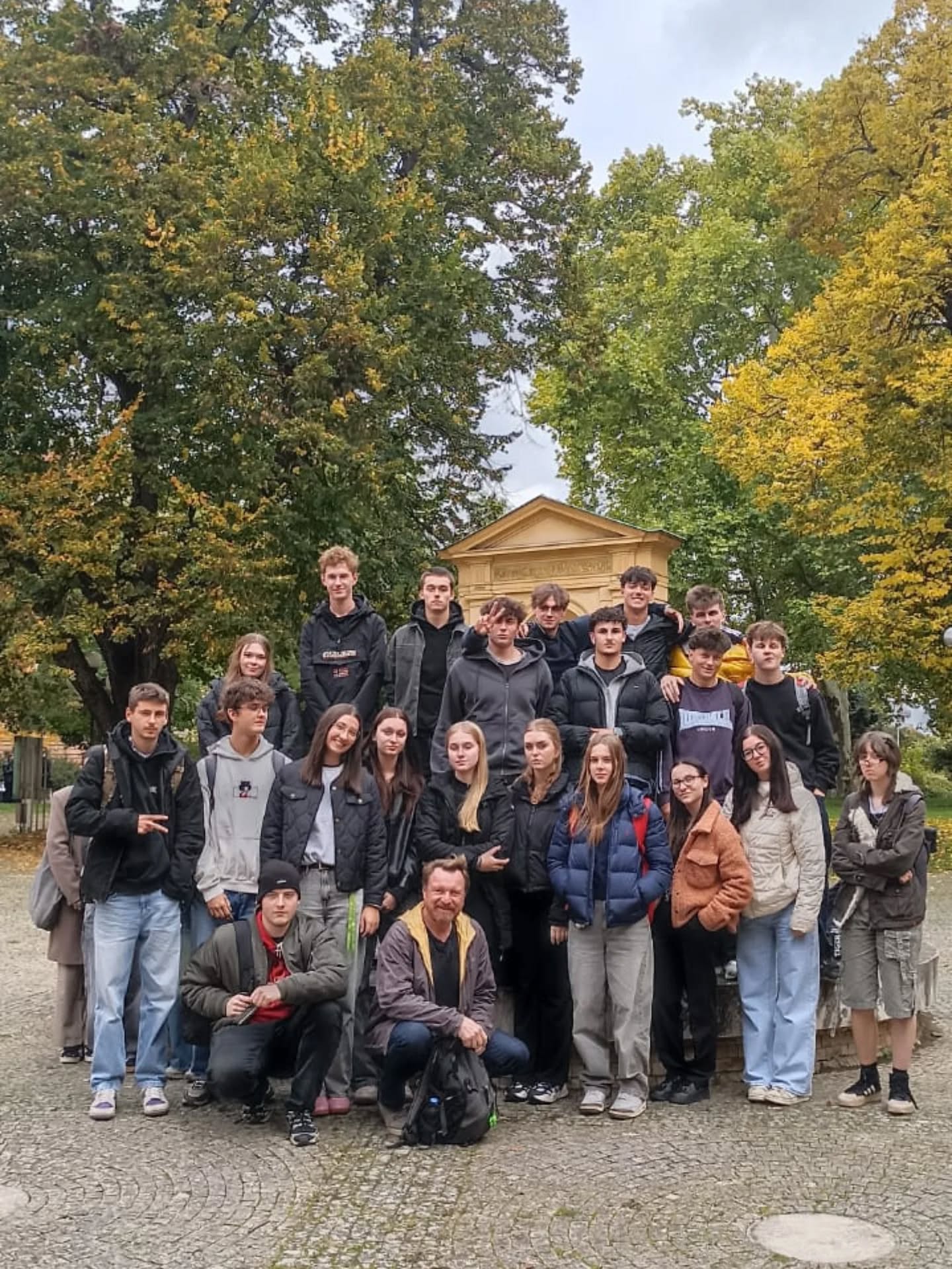 Skupina studentů s pedagogem pózuje na procházce v podzimním parku před historickým altánem.