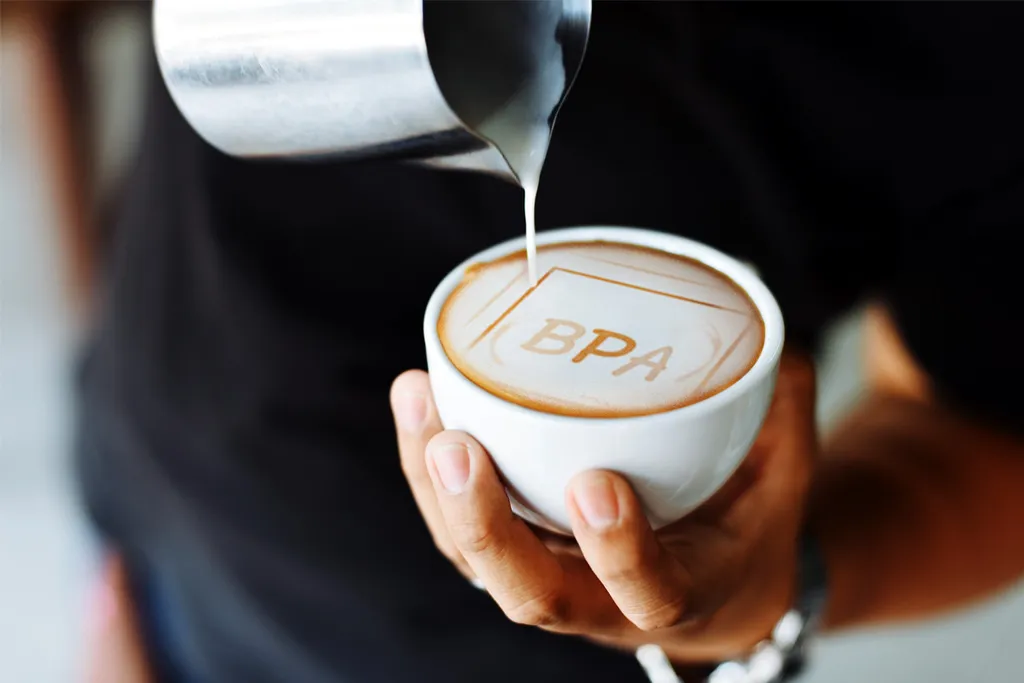 Barista nalévá mléko do kávy s motivem nápisu BPA ve pěně.