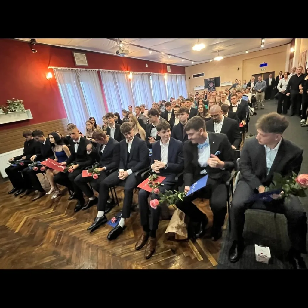 Absolventi v oblecích s růžemi sedí v sále během slavnostního předávání maturitních vysvědčení.