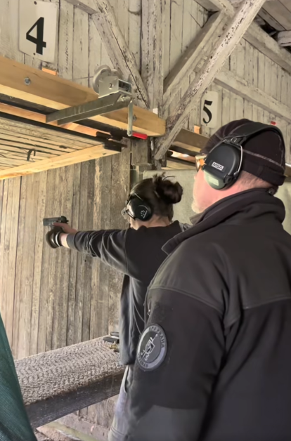Žákyně míří pistolí na střelnici pod dohledem instruktora, oba se sluchátky na ochranu sluchu.